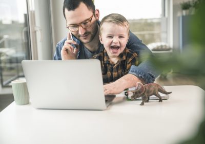 Vater mit Kind auf dem Schoß am Notebook und Handy als Metapher für "Mit Stress im Homeoffice entspannt umgehen"