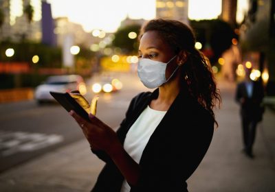 Business-Frau mit Mund-Nasen-Maske blickt auf Smartphone-Display als Metapher für den "zeb.market flash Q2 2020 (Issue 33 – Juli 2020)"